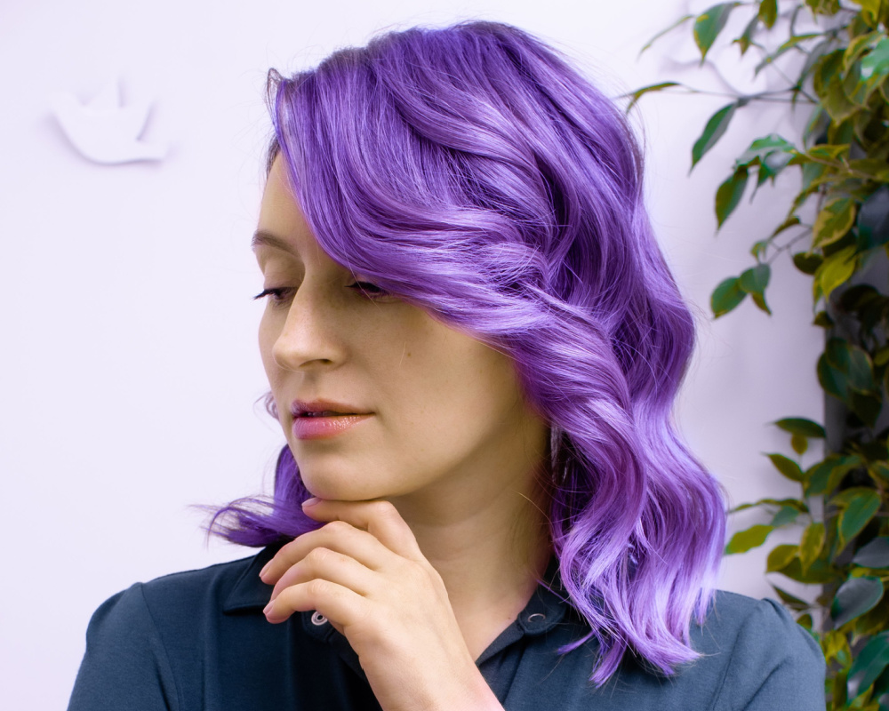 Close up of woman with hand rested under her chin looking down and away with wavy shoulder-length lavender hair and green leaves against the wall behind her