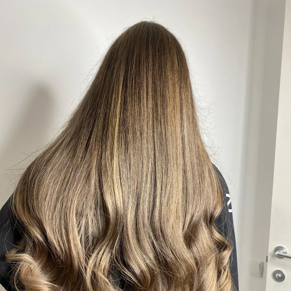 Back view of woman with long, curled hair featuring the popular dirty blonde hair color in front of a neutral wall