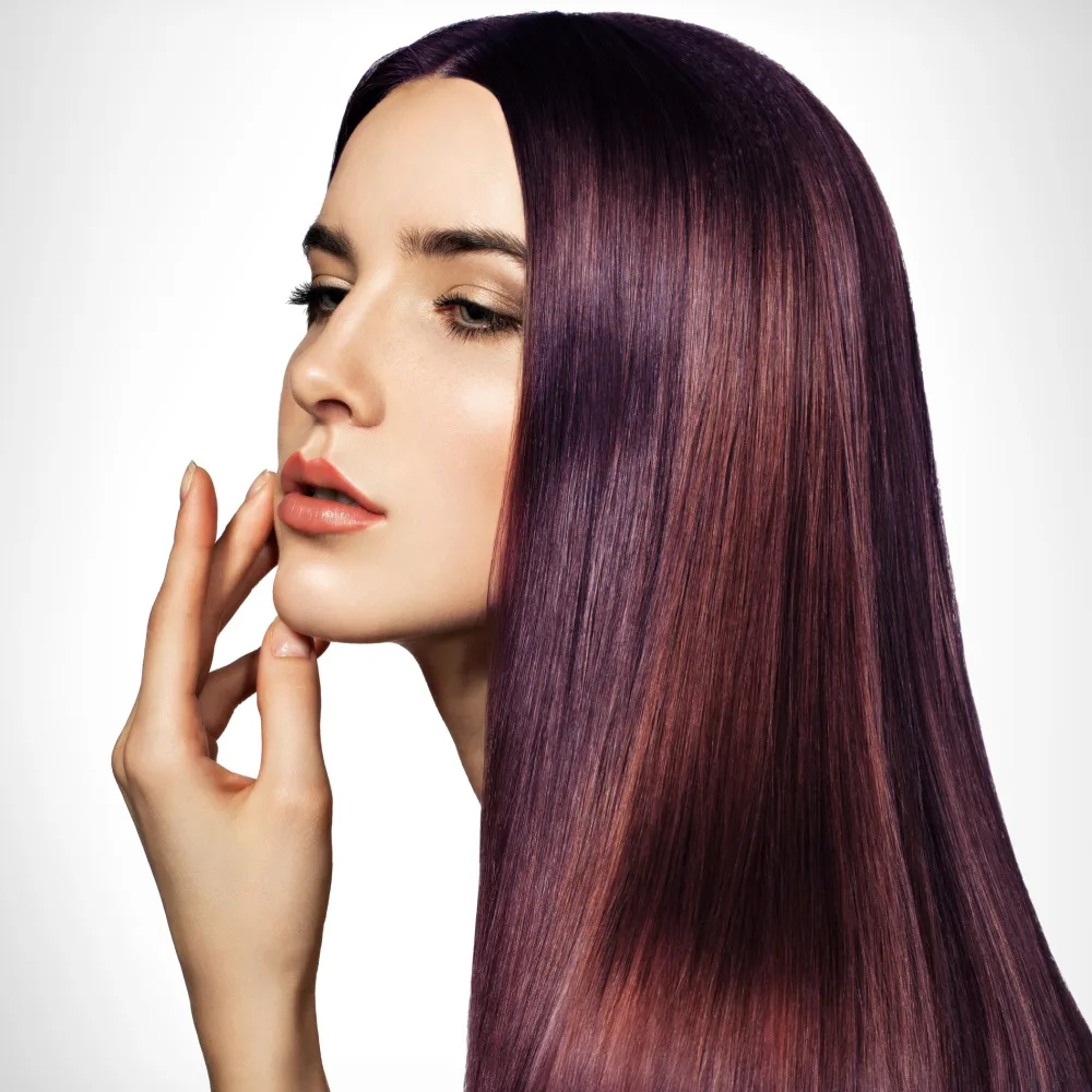 Model with long straight plum colored hair touches her chin facing the side in front of a white wall