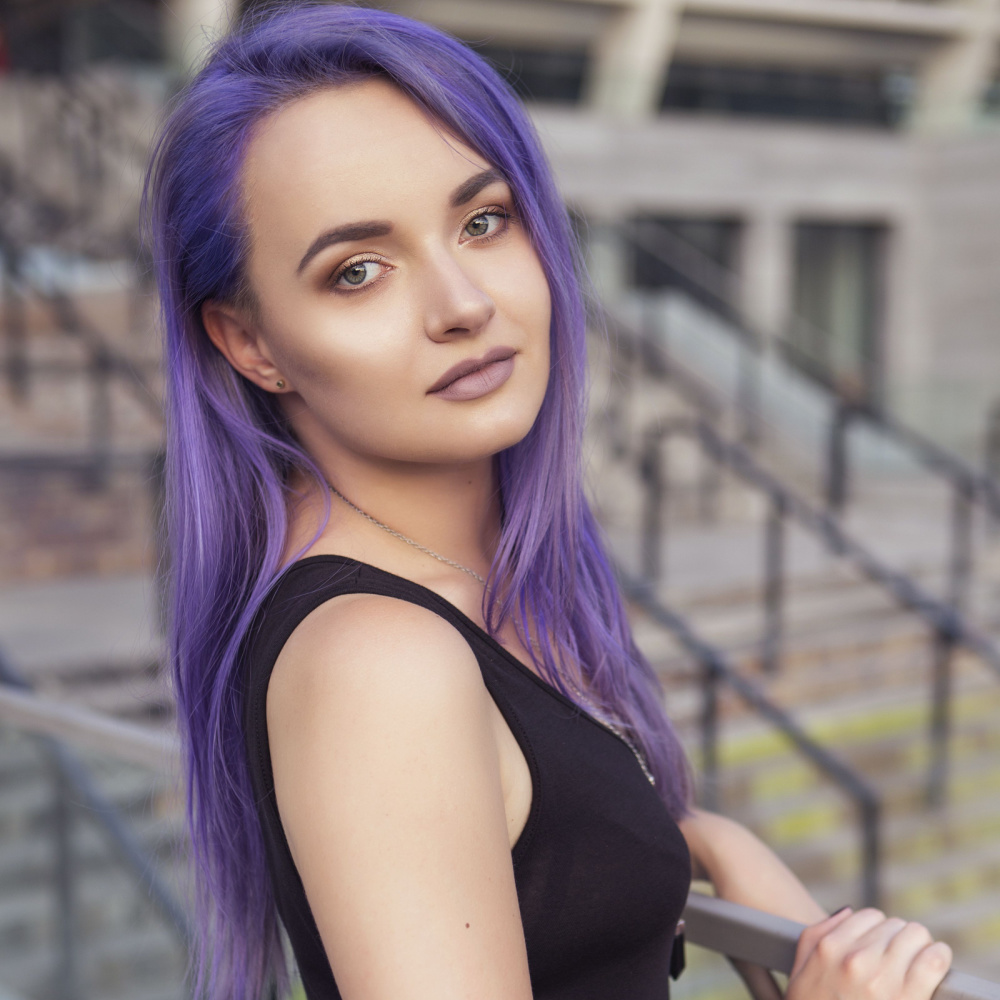Woman with black tank top stands at a railing in the city with her straight purple long hair styled with a side part
