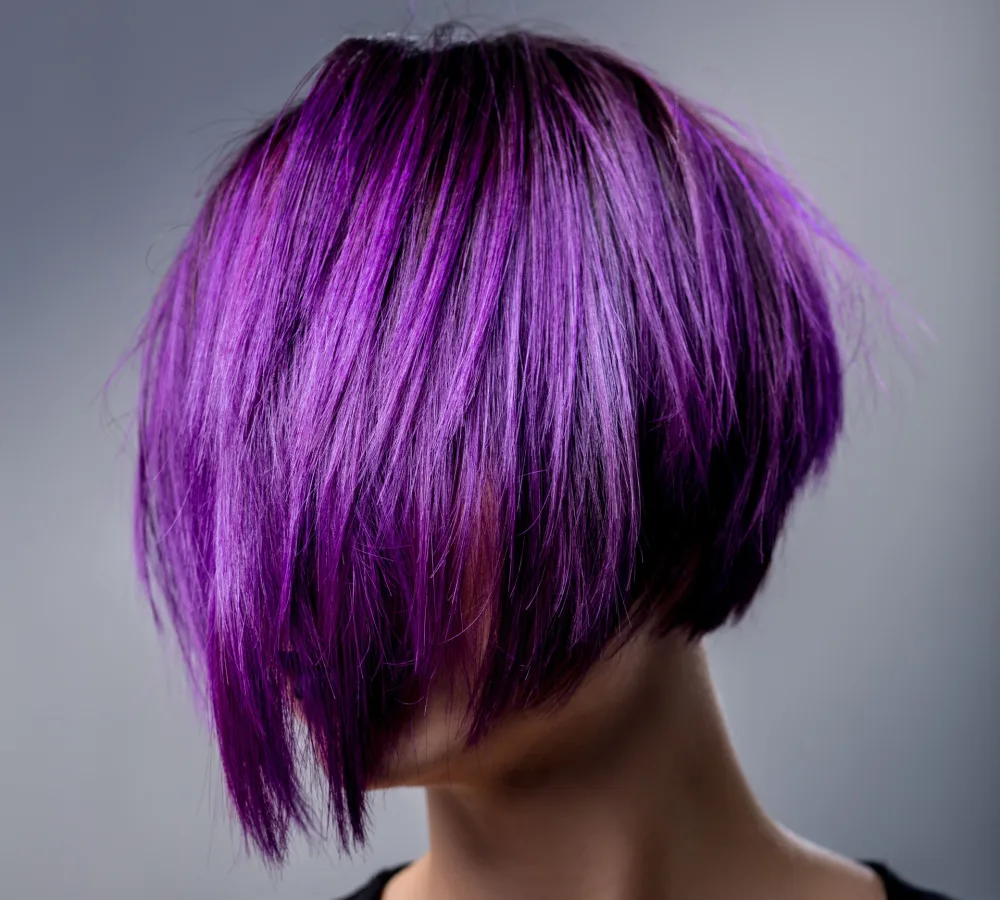 Woman with violet hair brushed down over her face shows off a short pixie bob haircut with vivid color in front of gray background