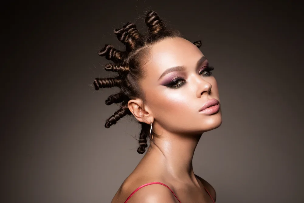 Woman wears Bantu knots as an example of cute black hairstyles with strong eye makeup in front of a gray dark wall