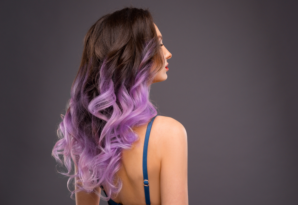 Back side view of woman with long brunette hair and lavender balayage color in a spaghetti strap top in front of charcoal wall