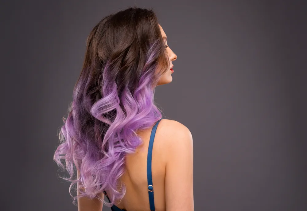 Back side view of woman with long brunette hair and lavender balayage color in a spaghetti strap top in front of charcoal wall