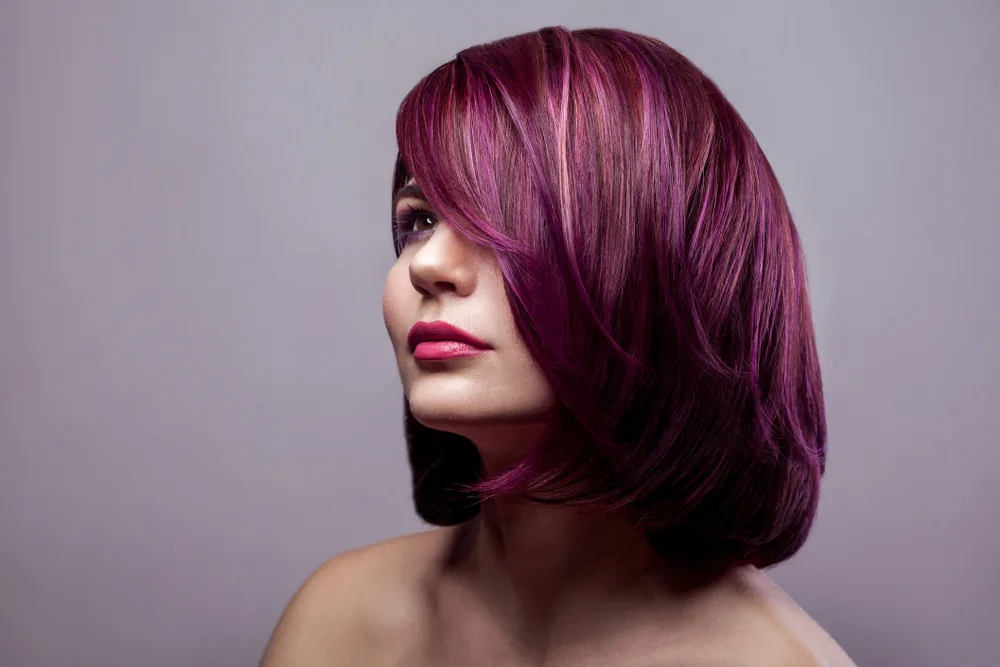 Front side view of woman with shoulder length plum colored bob with subtle highlights in front of gray wall