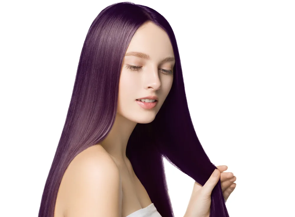 Fair woman with long straight plum colored hair holds out a section of hair in front of a white background