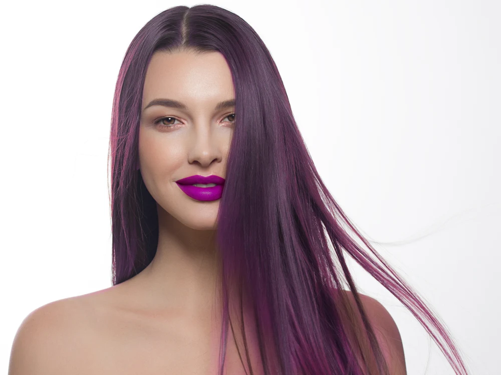 Woman with bare shoulders and long straight plum colored hair holds out a few strands with bright magenta lipstick on