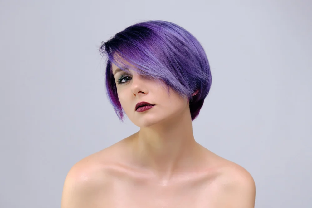 Woman with bare shoulders models her lavender color with a bixie cut and lighter purple highlights in front a gray wall