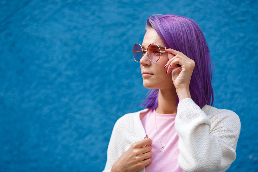 Woman in white jacket and pink top wears big round sunglasses with her bright lavender lob hairstyle in front of a blue wall