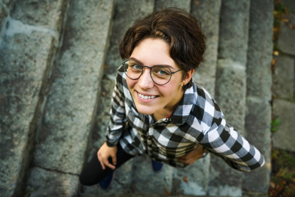 Pretty tomboy in a plaid checkered shirt and round glasses smiling and looking up at the camera for a piece on the best tomboy short fluffy haircuts