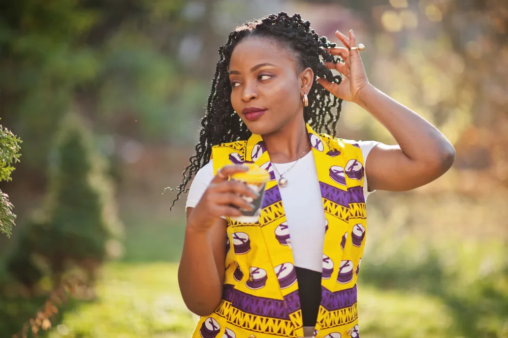 Woman of color poses with hand tucking her spring twist hair behind her ear outside holding a glass of water in a yellow and purple patterned dress