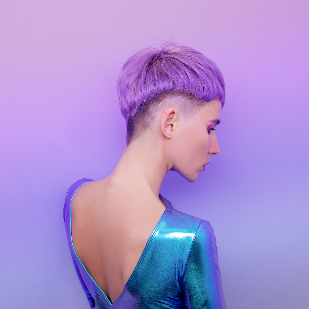 Back view of woman with short lavender undercut hairstyle wears a shiny blue leotard in front of gradient purple background