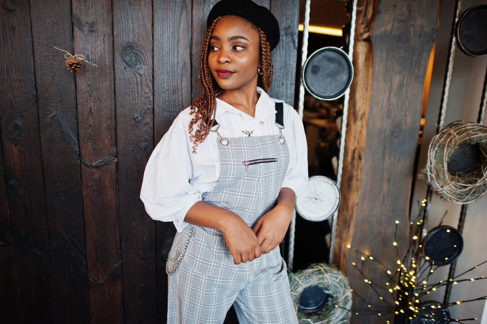 Black woman in overalls and white button down shirt looks to the side wearing spring twists and a hat in front of wooden wall