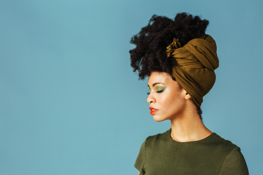 Mixed race woman with closed eyes and bright makeup wears a hair wrap style with a green color scheme in front of a blue wall