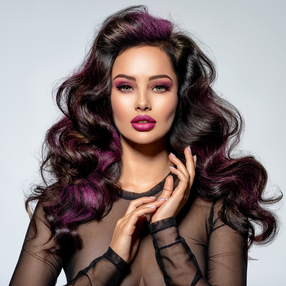 Woman wears a mesh black top with her long brown curly hair accented with purple plum color