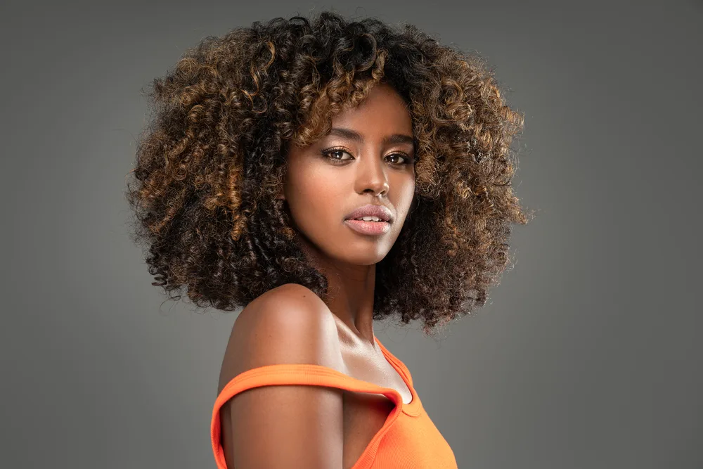 African American woman with orange tank top wears an example of cute black hairstyles with loose coils and balayage color