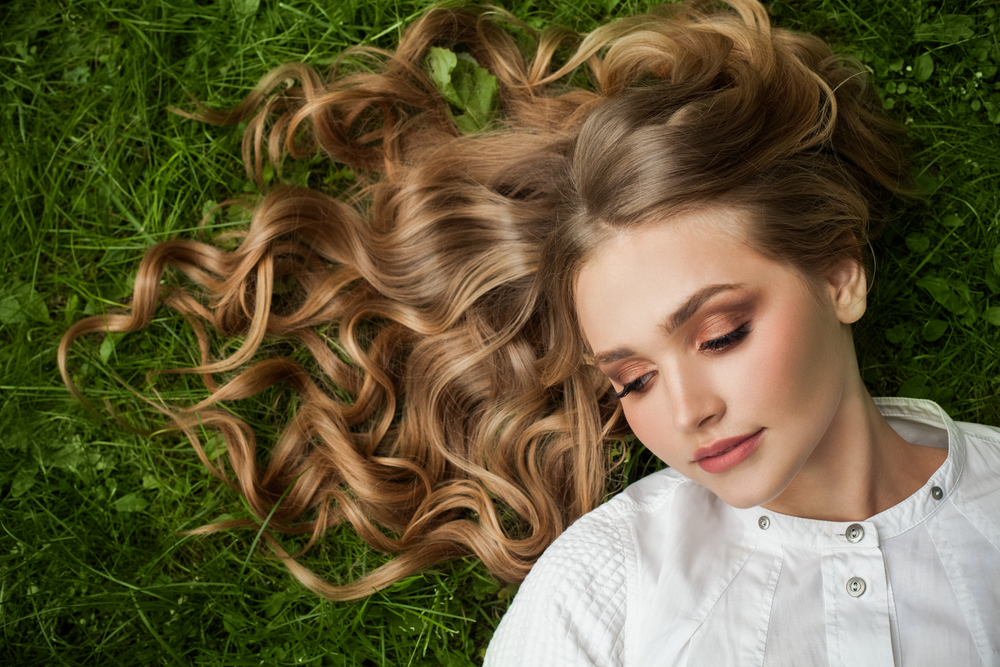 Young woman lays on grass with curly hair spread around her wearing a white shirt