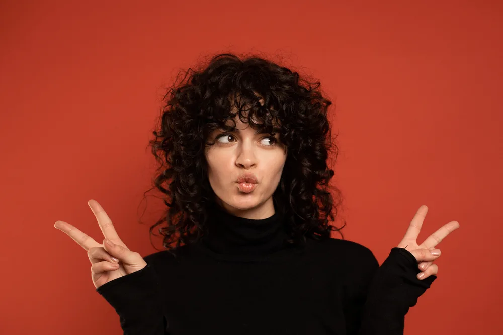 Curly Shag With Wispy Bangs on a woman with an 80's vibe giving the peace sign in a red room
