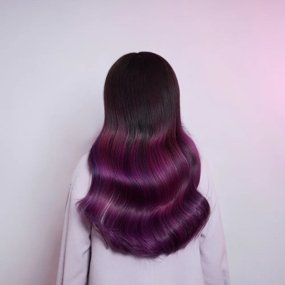 Back view of plum balayage hair color with magenta tones in front of pastel wall