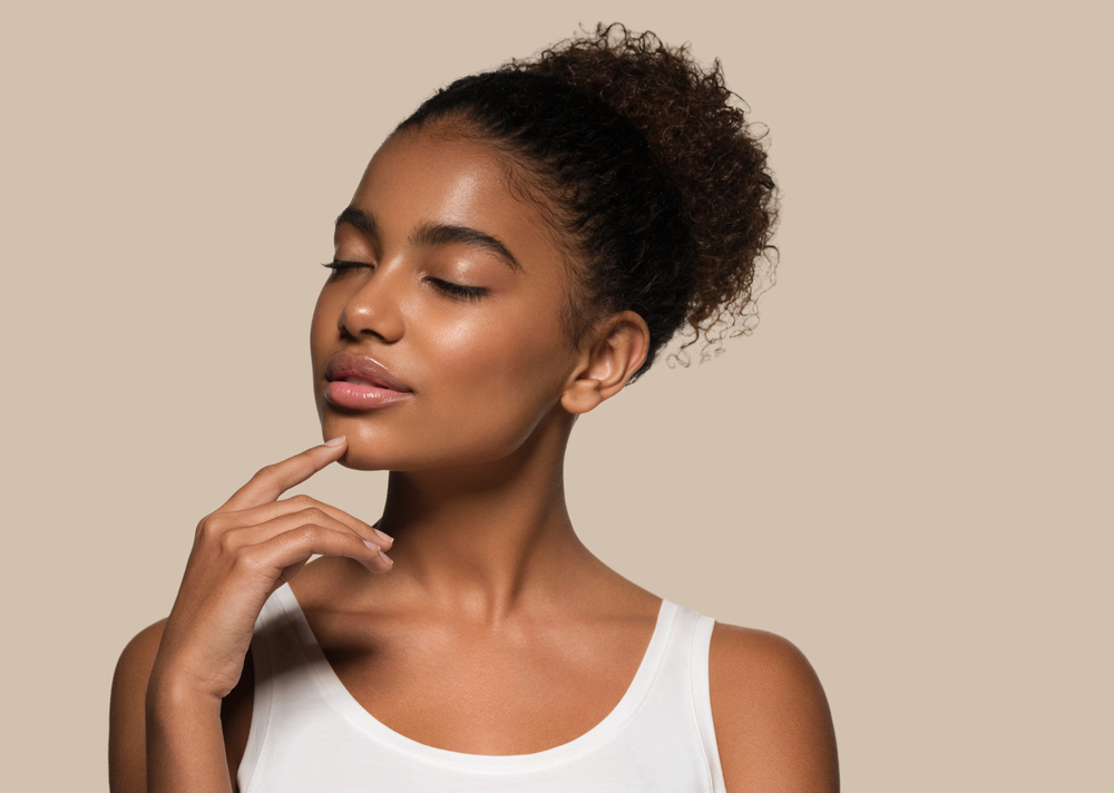 Black woman closes her eyes and touches her chin with white tank top while wearing a puffy ponytail hairstyle