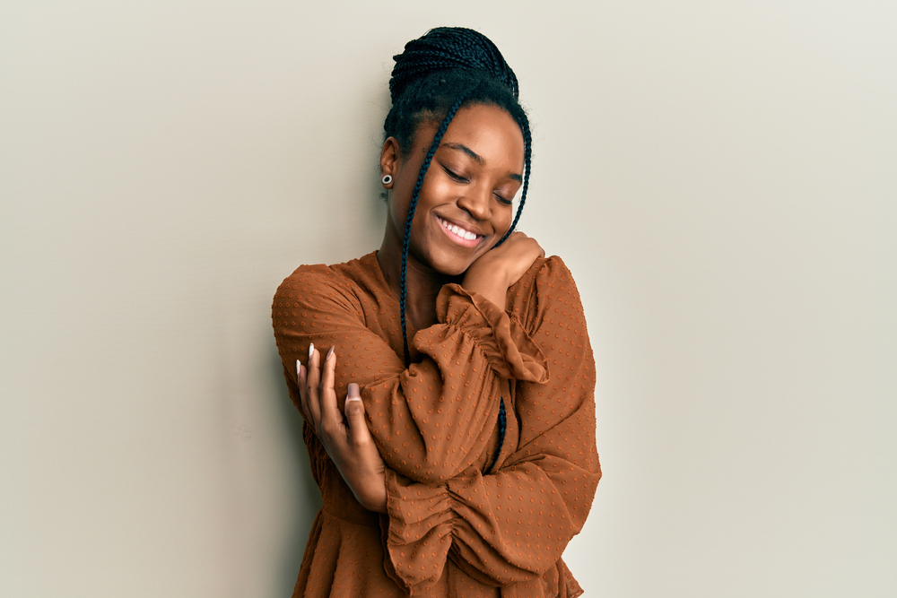 Young black woman hugs herself and shows off her favorite cute black hairstyle with a braided bun and face-framing braids hanging in front with a brown long sleeved shirt 
