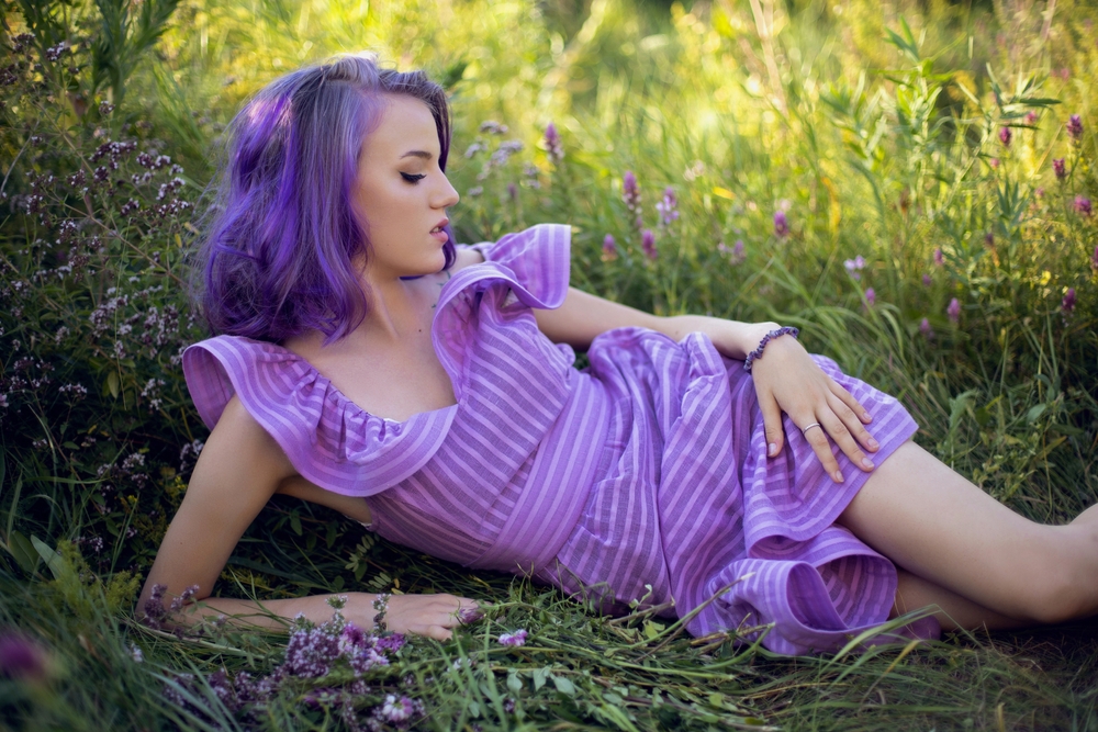 Woman lies in tall grass with purple dress and matching hair while looking away