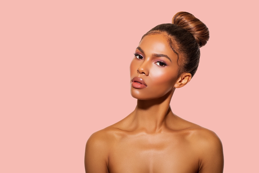Woman with dark tan skin and bare shoulders wears a cute black hairstyle for relaxed hair with a high ballerina bun in front of a pink wall