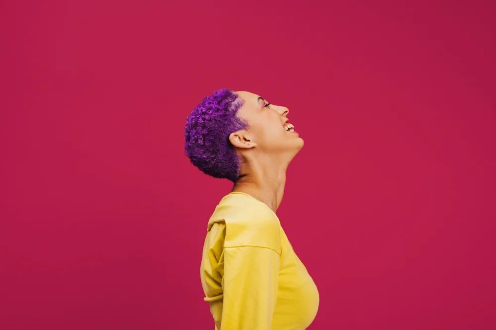 Woman with lavender hair and medium skin tone laughs with her head raised and eyes closed while wearing a yellow shirt in front of a red background