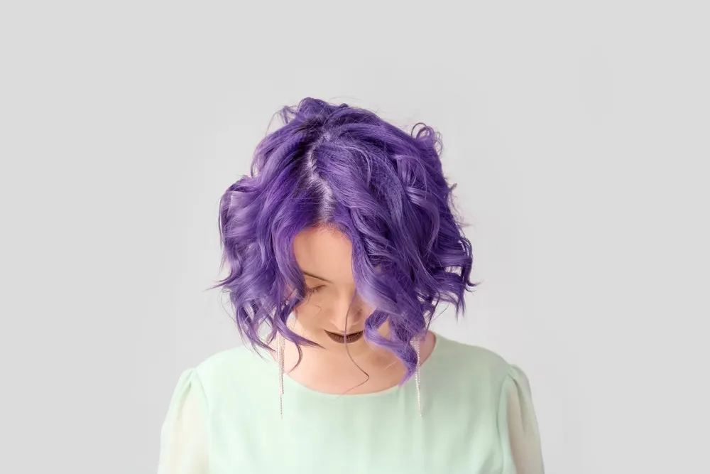 Woman looks down with lavender hair styled in a short curly bob with a pastel green shirt in front of gray wall