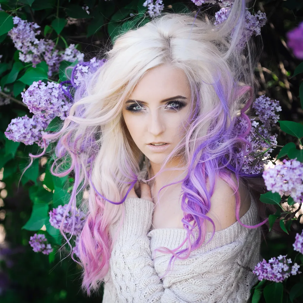 Blonde woman stands in front of flower bushes with pink and lavender hair colors and white off-shoulder sweater