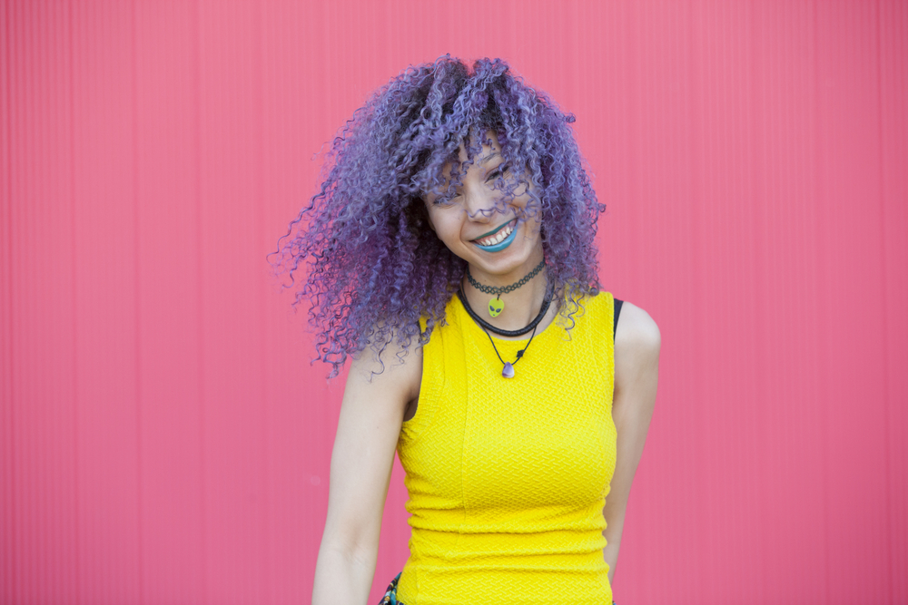 Light-skinned woman with lavender coily hair smiles and wears a yellow tank top in front of a pink wall