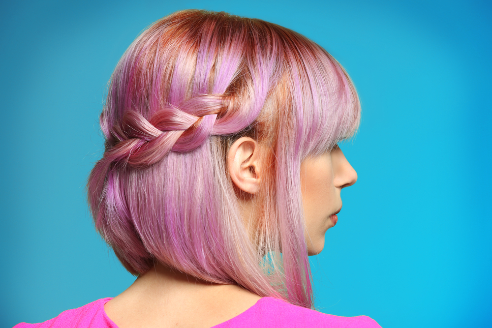 Young woman with purple hair color wears a half up braid hairstyle perfect for prom in front of blue wall