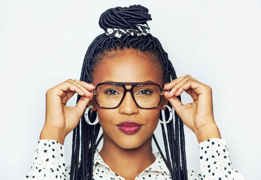 Woman puts on dark rimmed glasses with her faux locs and topknot hairstyle wearing a white polka dot shirt and hoop earrings 
