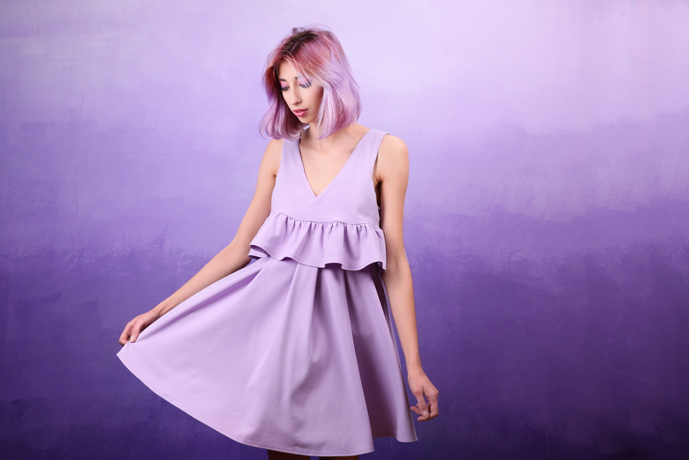 Girl with purple hair and ruffled purple dress looks down holding the hem of her dress in front of purple gradient background
