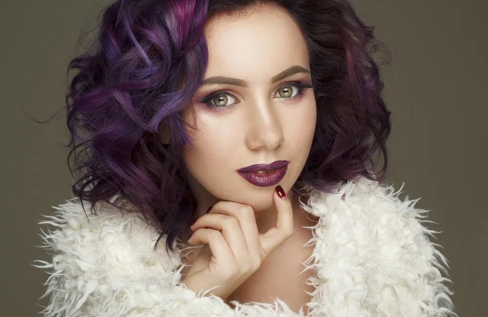 Model with white furry top touches her chin with a short plum and violet colored bob hairstyle
