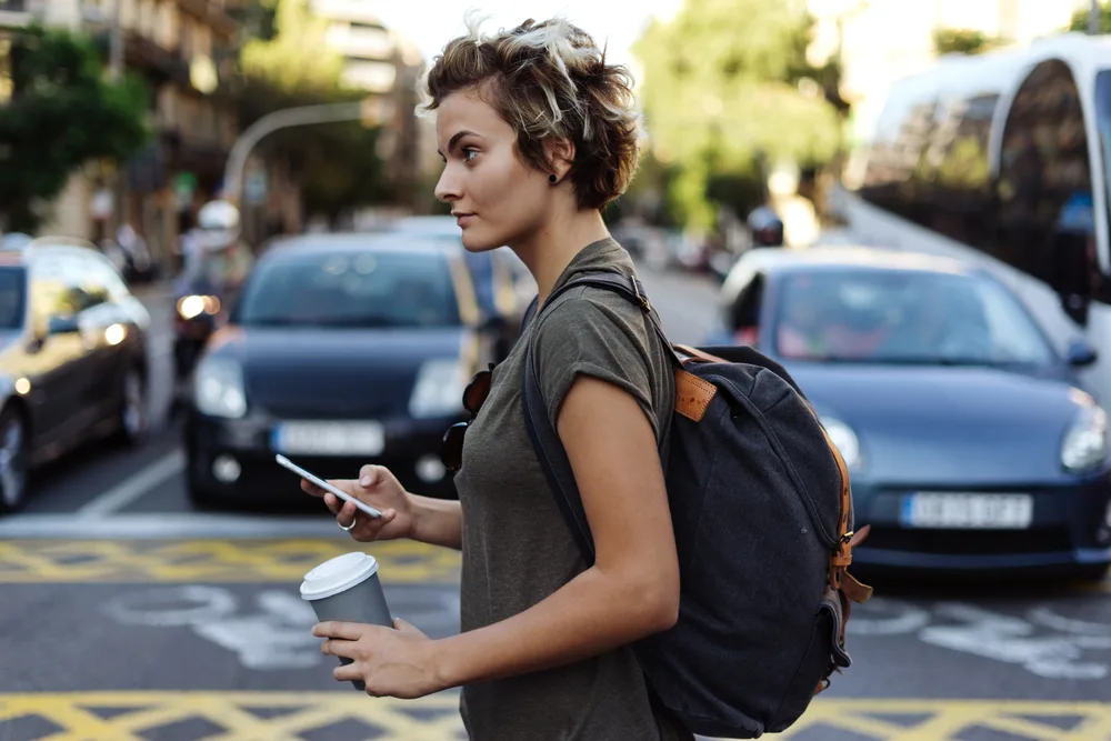 Tomboy walking in a city with a coffee in her hand, a short fluffy haircut on her head, and a backpack on her back