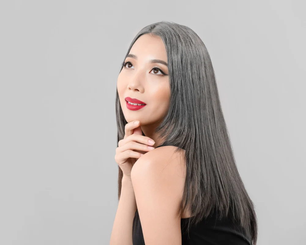 Asian woman with straight, layered reverse grey ombre hair color smiles with her hand thoughtfully posed under her chin in front of a grey wall