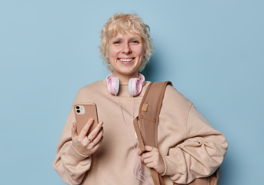 Blonde woman smiles holding a smartphone with a brown backpack over her shoulder and headphones around her neck wearing a short blonde wavy haircut that suits oval faces
