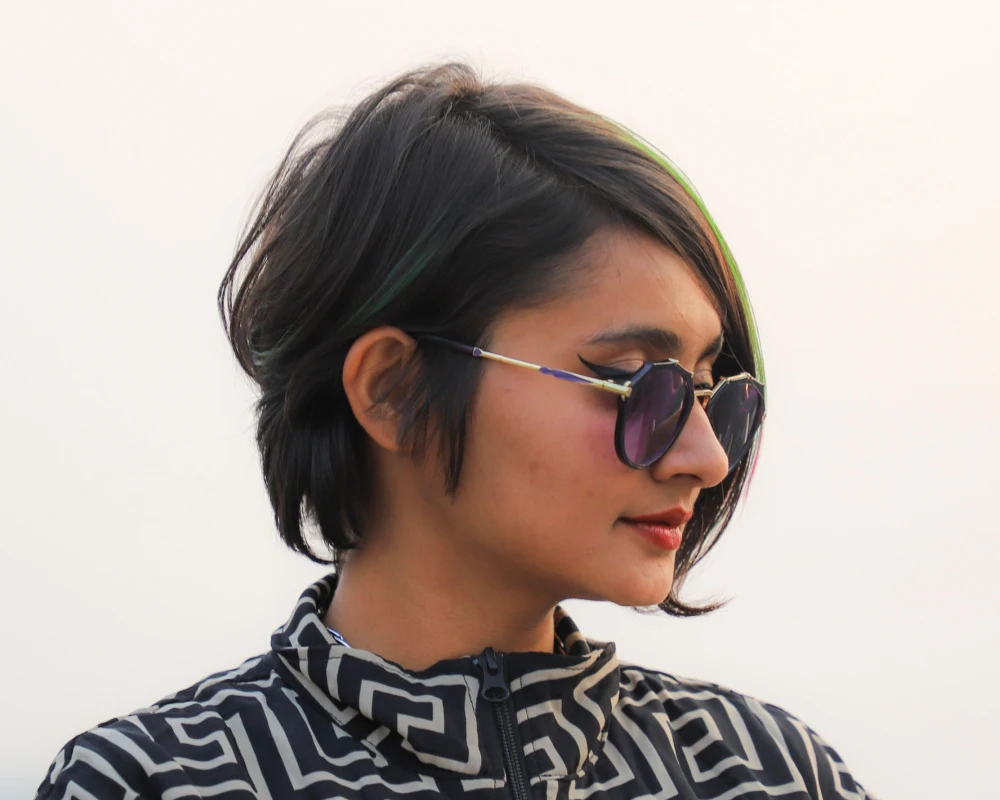 Woman with short brown hair wears sunglasses and a textured bixie cut parted to the side as one of the best short haircuts for oval faces while glancing down and away