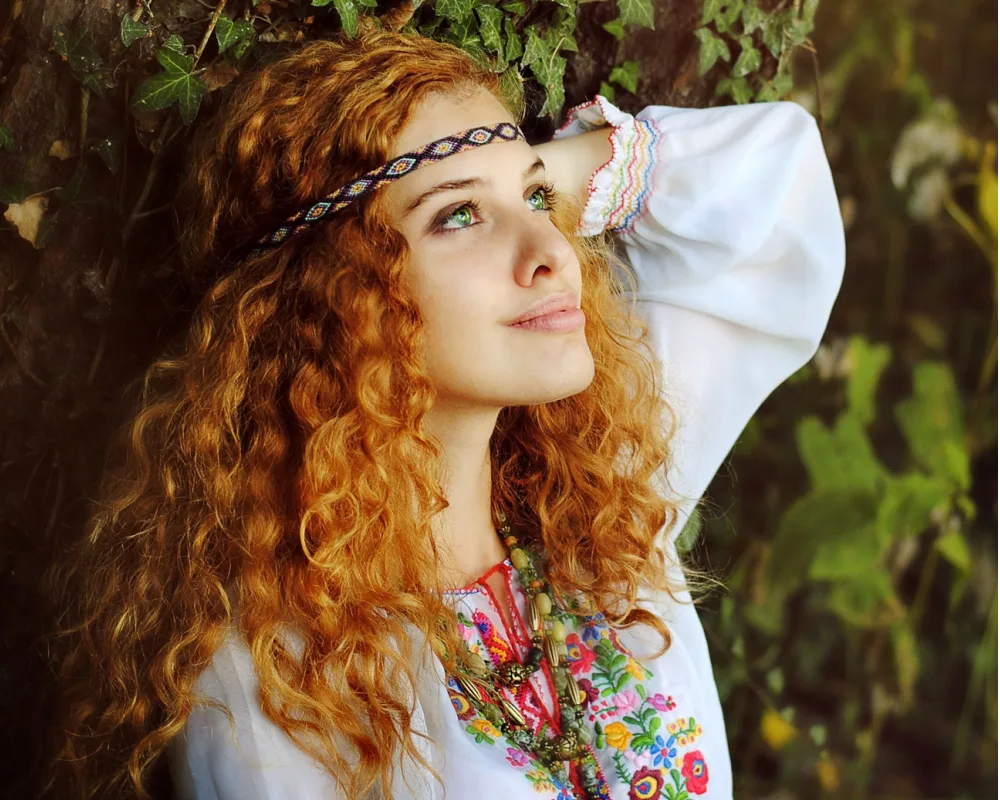 Woman with copper red hair looks up smiling in a forest with curly hair and a boho headband across her forehead