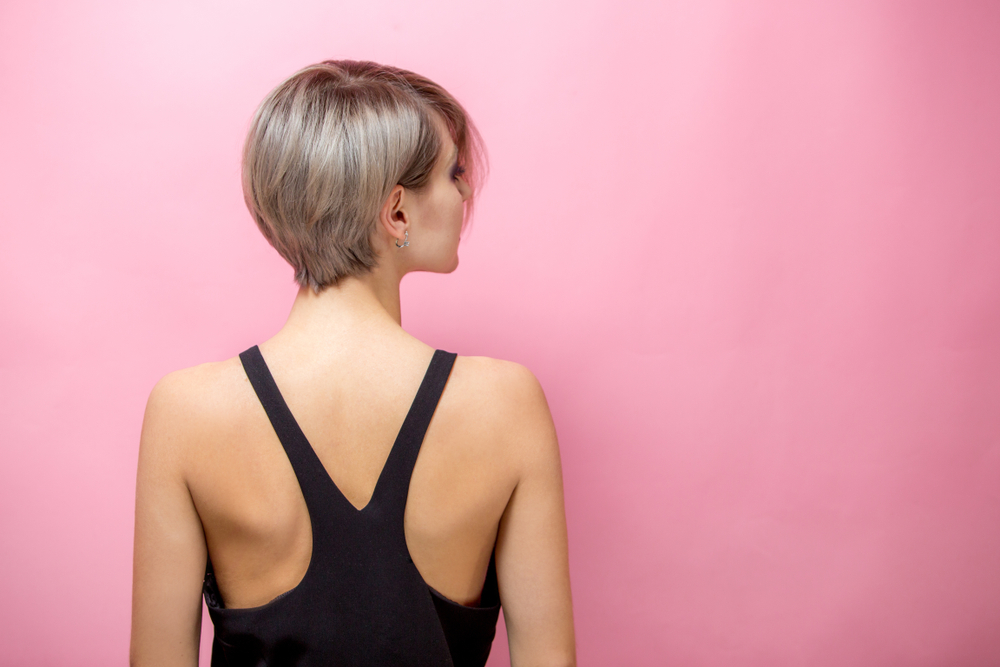 Rear view of woman with short pixie haircut facing a pink wall with a black racer back tank top on