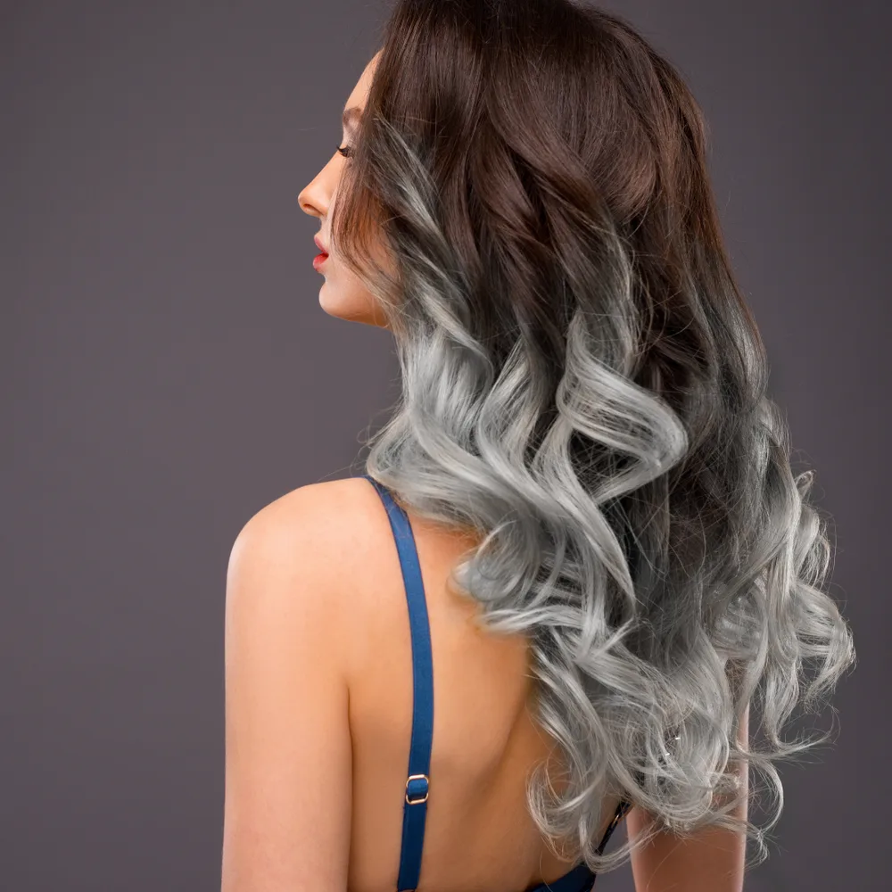 Side rear view of woman in a tank top with long, curly brown hair dyed grey at the ends while standing in front of dark grey wall