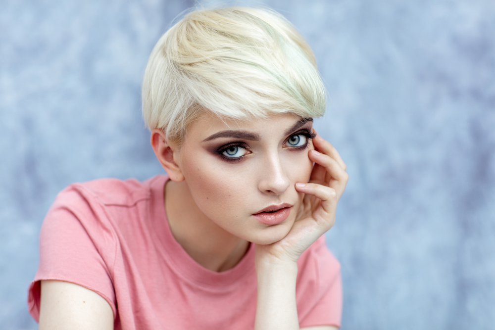 Example of short haircuts for oval faces include a messy pixie with side swept bangs, shown on a blonde model with a pink t-shirt