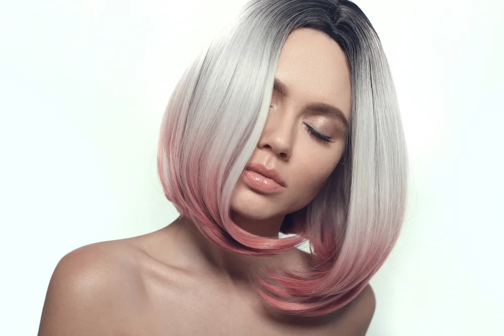 Woman tilts her head with bare shoulders posing in front of a white wall with grey ombre hair featuring pink colored ends