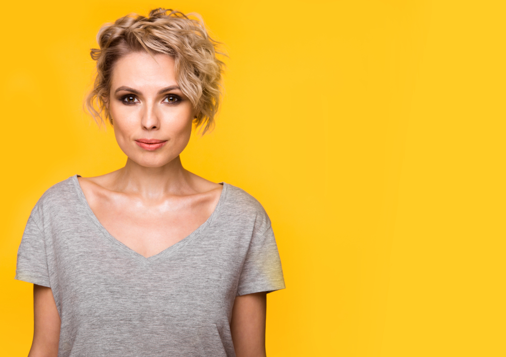 Blonde woman with wavy hair and side bangs wears a gray t-shirt in front of a yellow background to show off one of the best short haircuts for oval faces