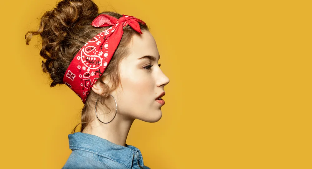 Woman with a curly bun wears a bandana tied around her head as a headband with a denim shirt in front of a yellow background