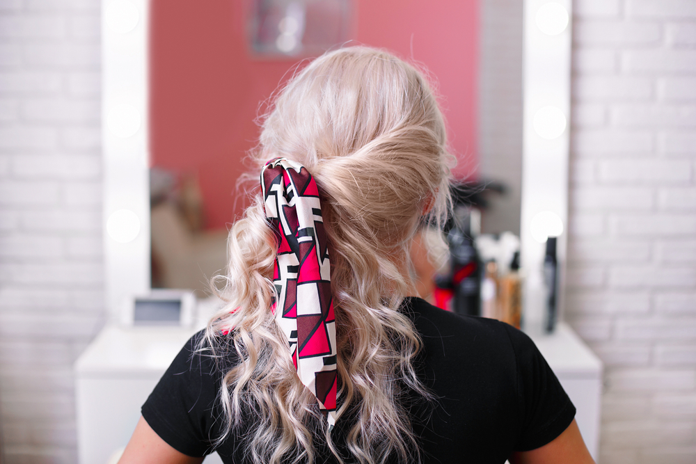 Rear view of a blonde woman in a salon wearing an inverted side ponytail with a scarf, an example of cute hairstyles for curly hair