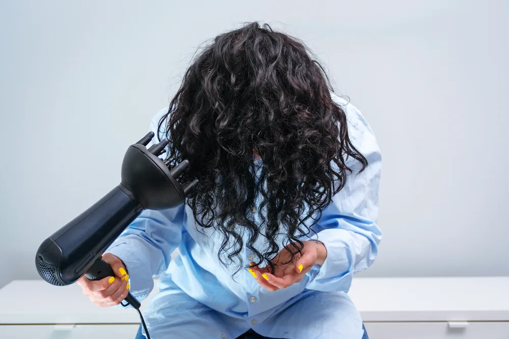 Woman sitting down bends forward to use a hair dryer with a diffuser attachment (a key tool to cute hairstyles for curly hair) to dry her hair after a shower