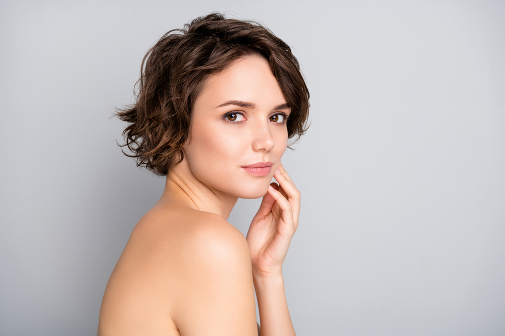 Brunette woman with a short bob haircut touches her cheek looking over her bare shoulder in front of a gray wall