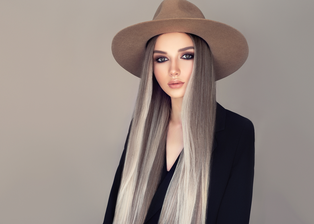 Woman with grey ombre hair color wears a brown hat and black long sleeved top with eye makeup in front of a light gray wall